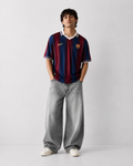 A man modeling a vintage-style FC Barcelona soccer jersey with red and blue stripes, paired with wide-leg gray jeans and white sneakers, standing against a light gray studio background.