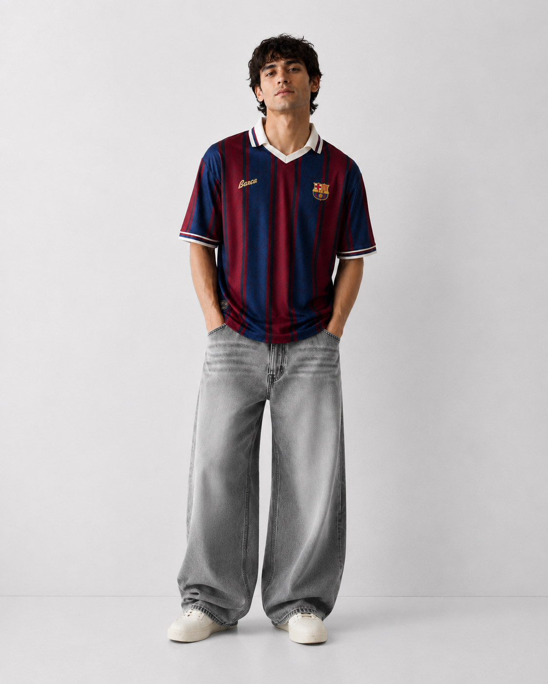 A man modeling a vintage-style FC Barcelona soccer jersey with red and blue stripes, paired with wide-leg gray jeans and white sneakers, standing against a light gray studio background.